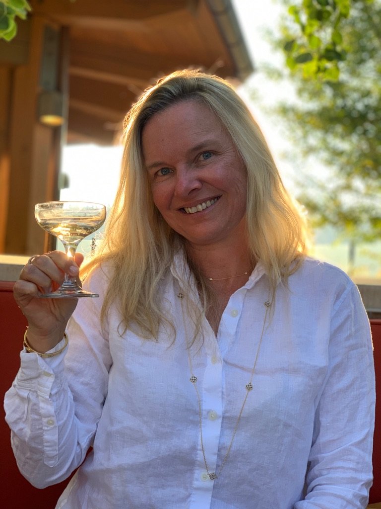 She-Stache in a white linen shirt raising a very small martini in a coupe glass on a sunlit patio