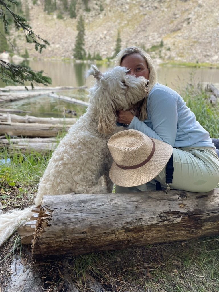 She-Stache sitting on a log by a mountain lake, hugging Pup-Stache, with her straw hat in her lap