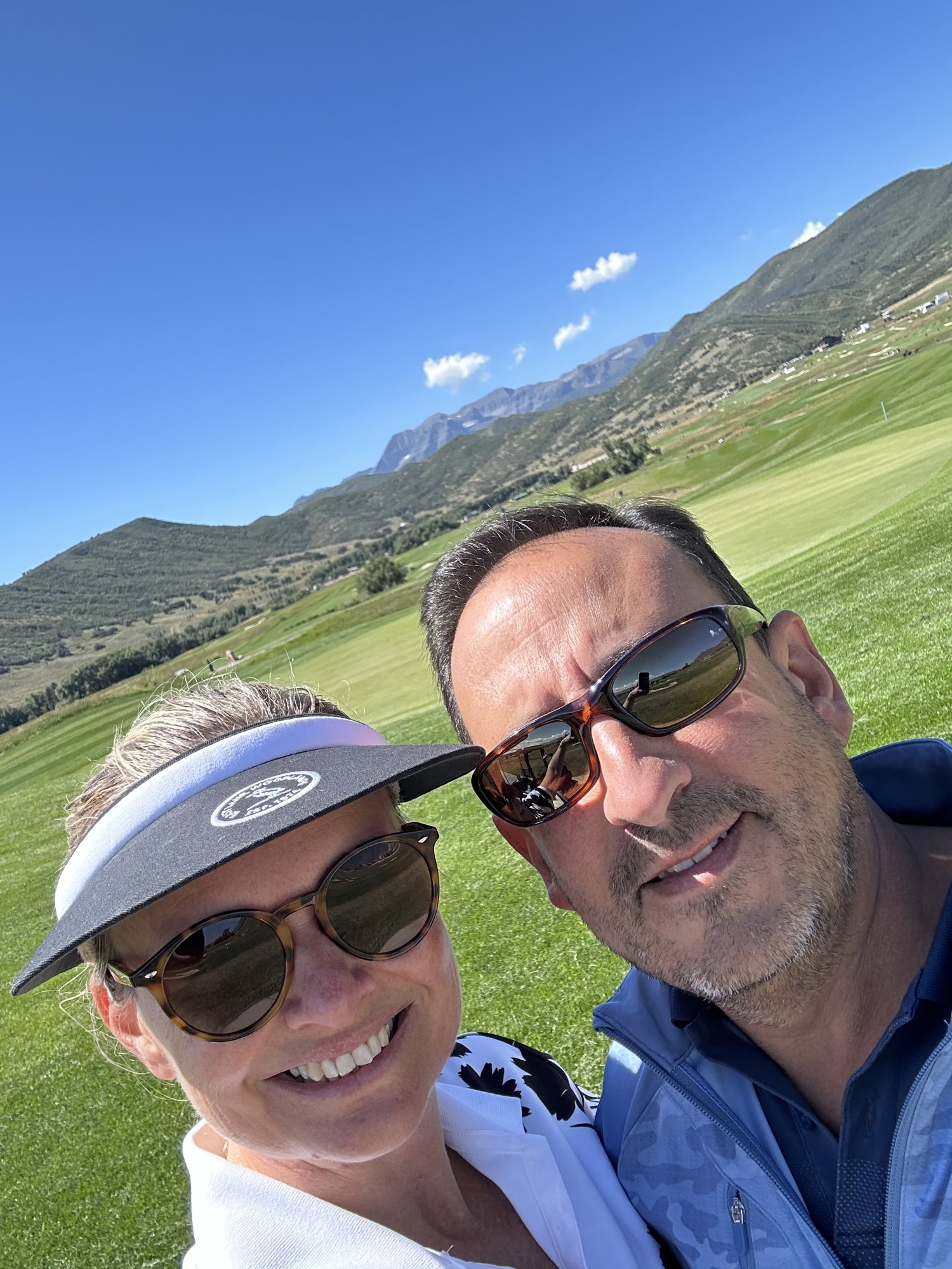 She-Stache in a white visor and He-Stache in sunglasses taking a selfie on a green fairway at Soldier Hollow with mountains behind them