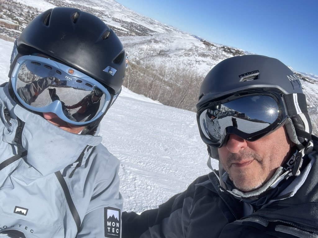 She-Stache and He-Stache in helmets and goggles taking a selfie on a groomed run, with the town below