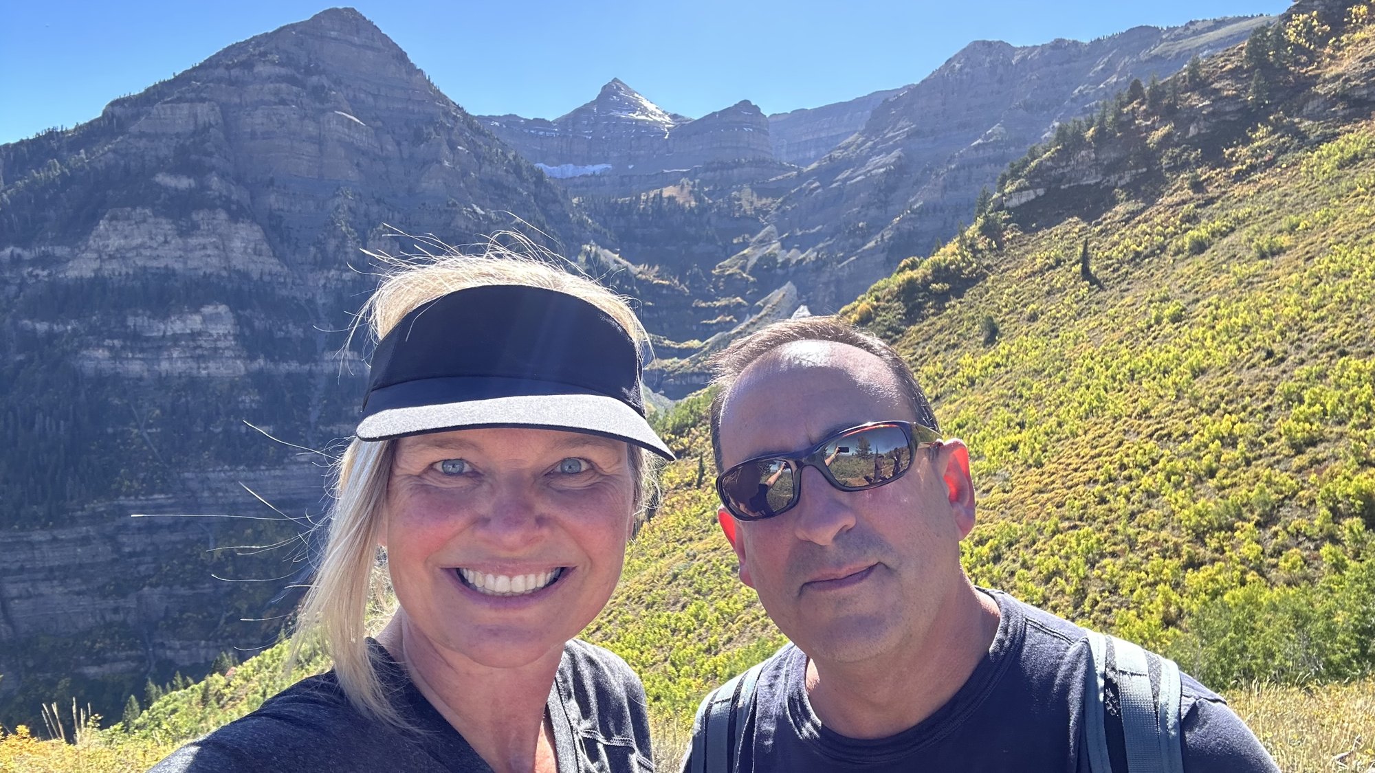 She-Stache in a black visor and He-Stache in sunglasses smiling on a green slope below the cliffs of Mount Timpanogos