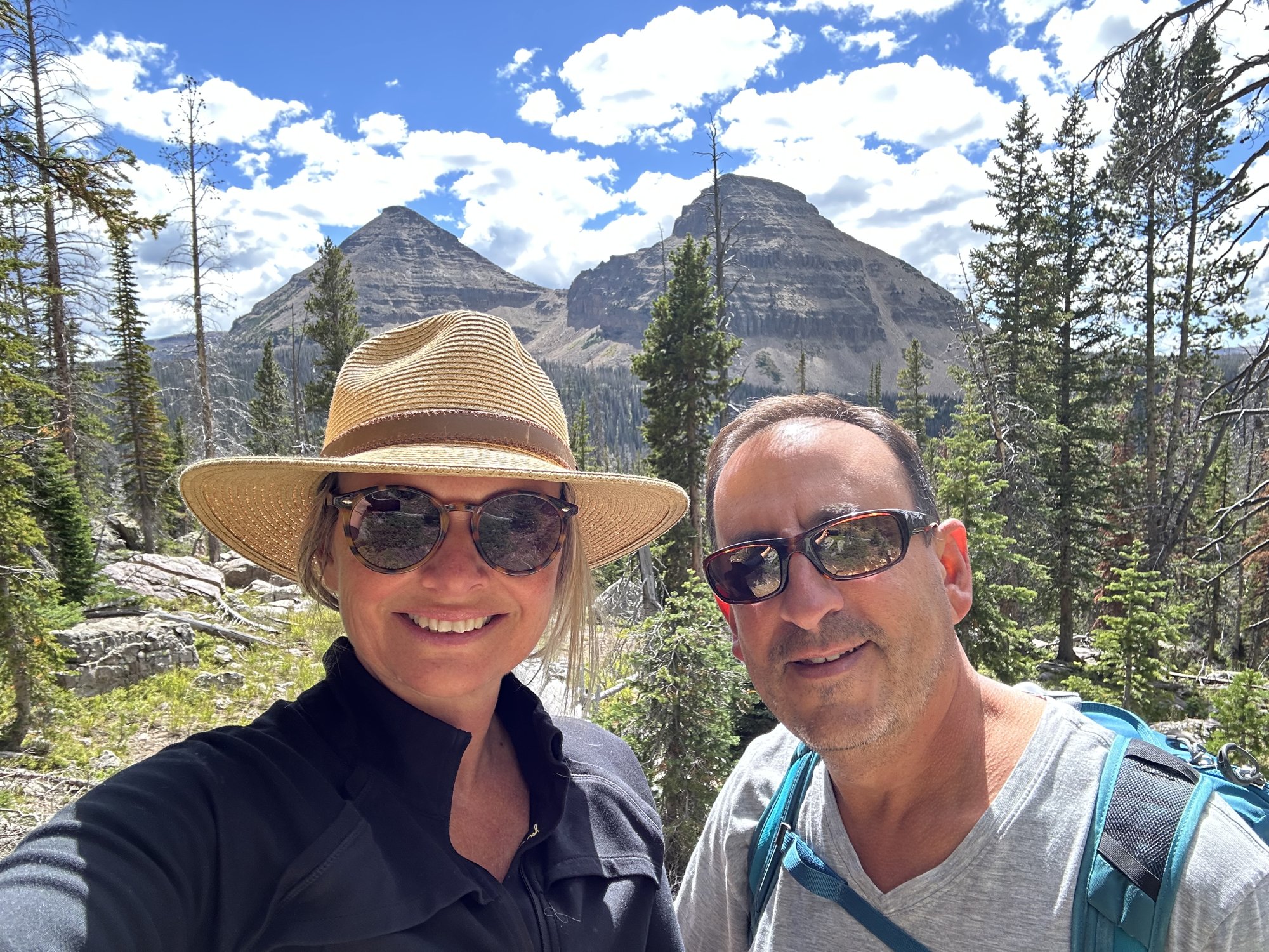 She-Stache in a straw hat and He-Stache in sunglasses taking a selfie in front of pine trees and rocky peaks in the Uintas