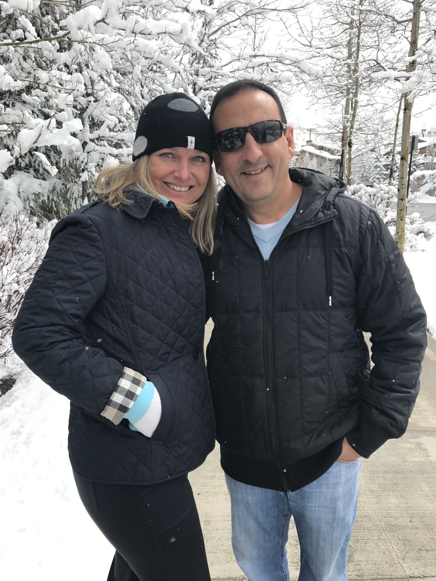 She-Stache and He-Stache in black winter jackets on a snowy sidewalk in Park City, with snow-covered aspens behind them