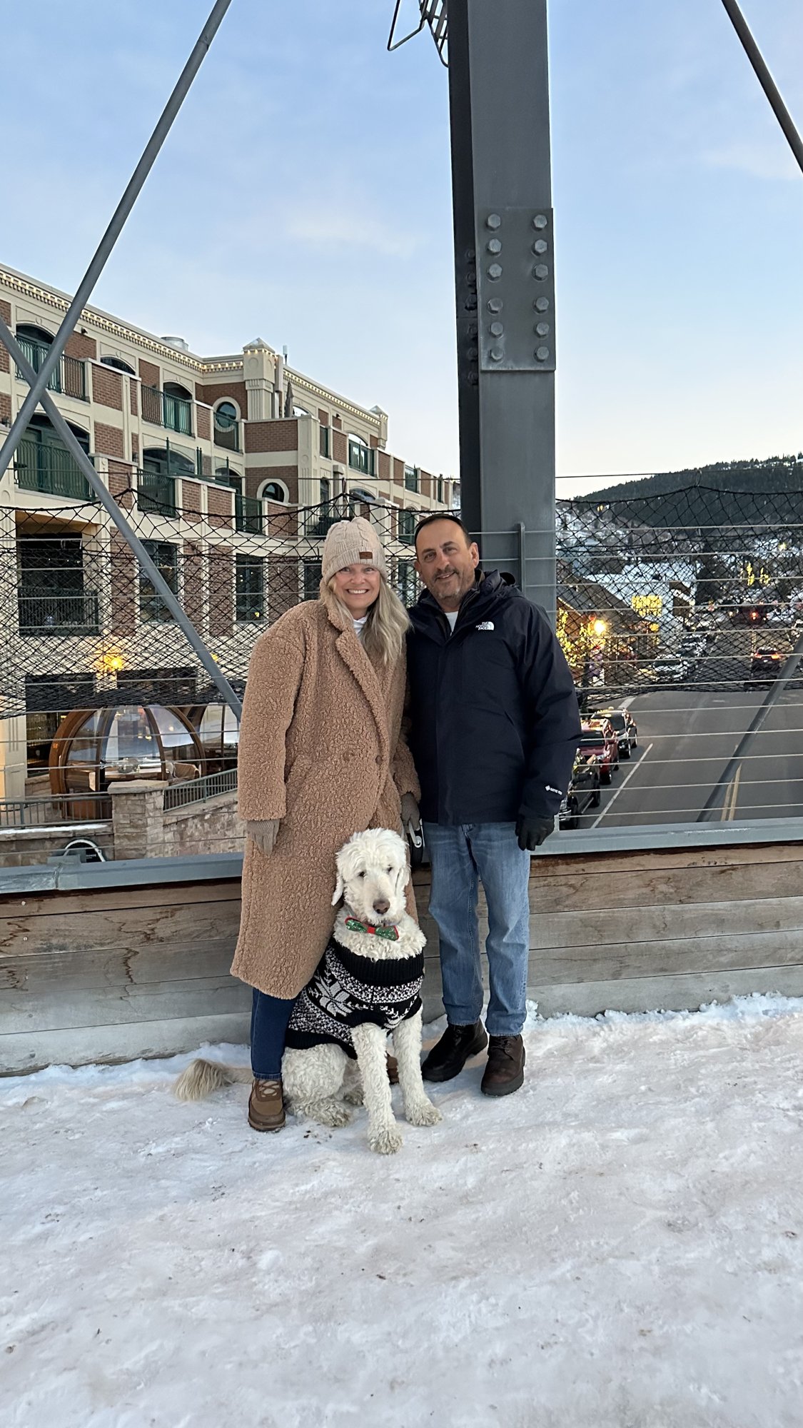 She-Stache in a camel coat and He-Stache in a black jacket on a snowy Main Street walkway, with Pup-Stache in a knit sweater sitting between them