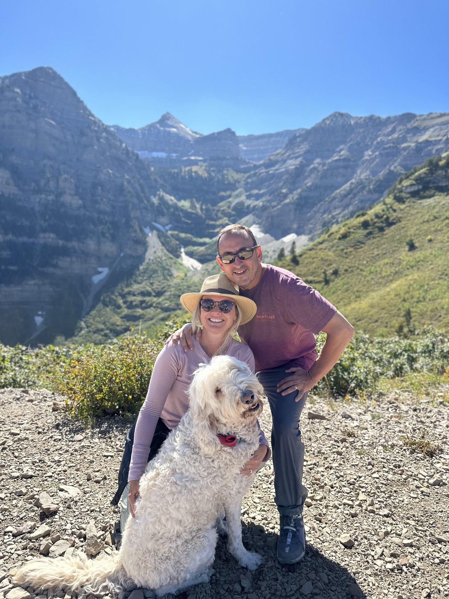 She-Stache, He-Stache and Pup-Stache posing on a rocky trail with a deep glacial cirque and gray cliffs of Mount Timpanogos behind them