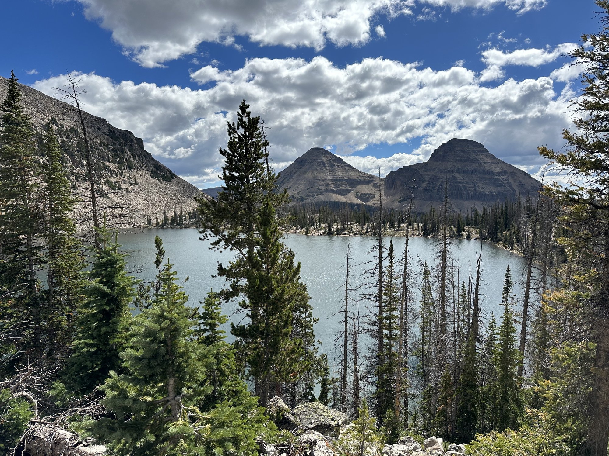 A blue alpine lake ringed by pine trees, with two rocky peaks and white clouds above, in the Uinta Mountains