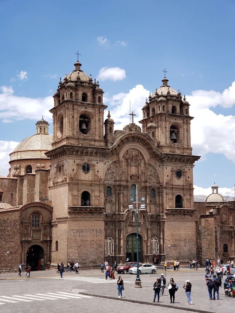 A brown stone baroque church facade with two bell towers and a dome under a blue sky, with people crossing a plaza in front