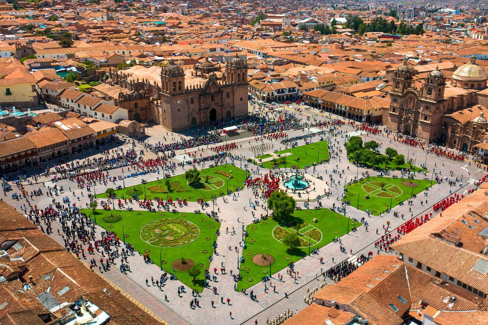 An aerial view of the Plaza de Armas in Cusco with green gardens, a fountain, crowds, and two baroque churches among red tile roofs