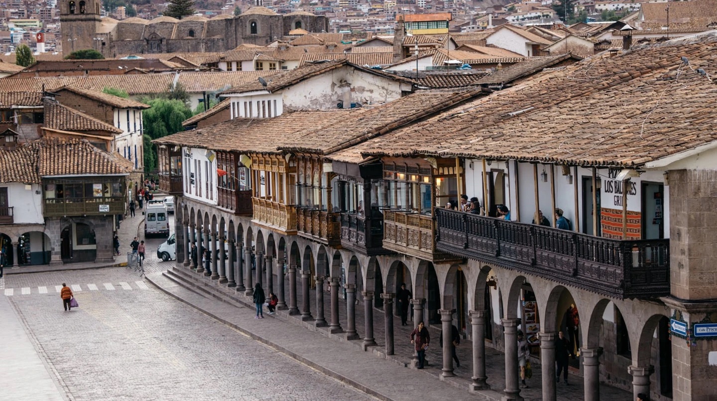 Carved wooden balconies over a stone arcade on a Cusco plaza, with tile roofs and a bell tower behind