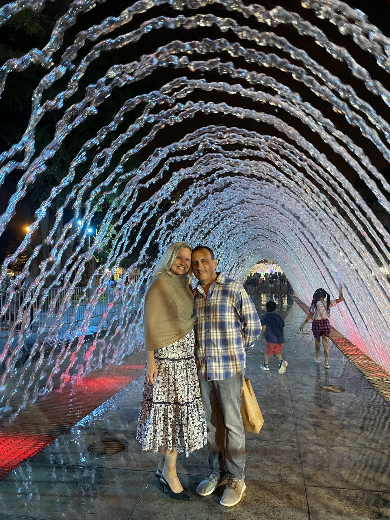 She-Stache and He-Stache inside a glowing tunnel of fountain arches at the Magic Water Circuit at night