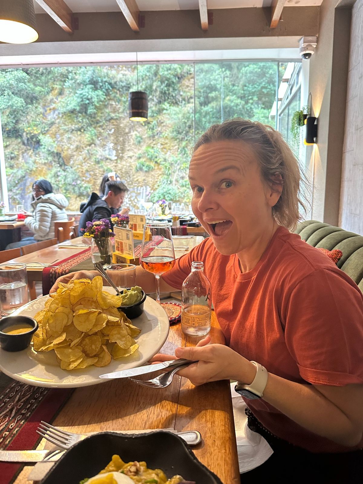 A woman in an orange shirt reacting to a plate of chips and guacamole at a restaurant table with a window onto the river