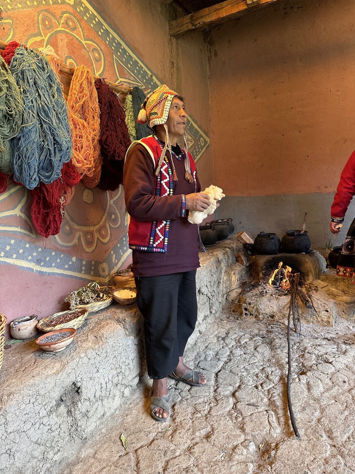 A weaver demonstrating natural dyes beside hanging skeins of colored yarn and a wood fire