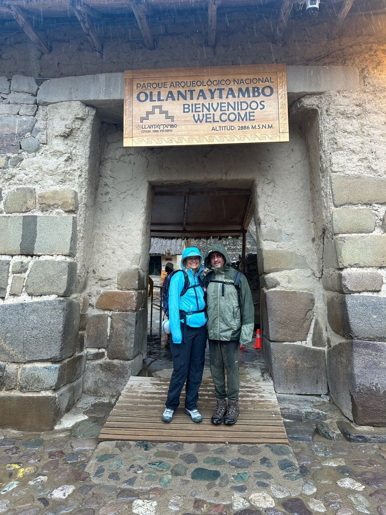 She-Stache and He-Stache in rain jackets under the Ollantaytambo archaeological park welcome sign