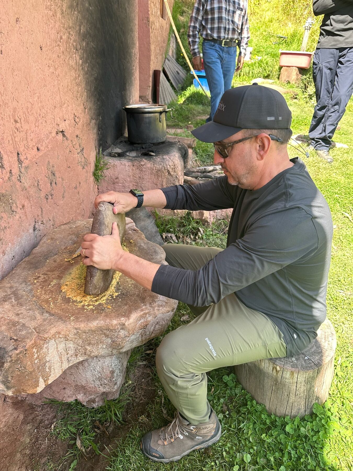 A man in a black cap grinding corn on a large stone mortar beside an adobe wall