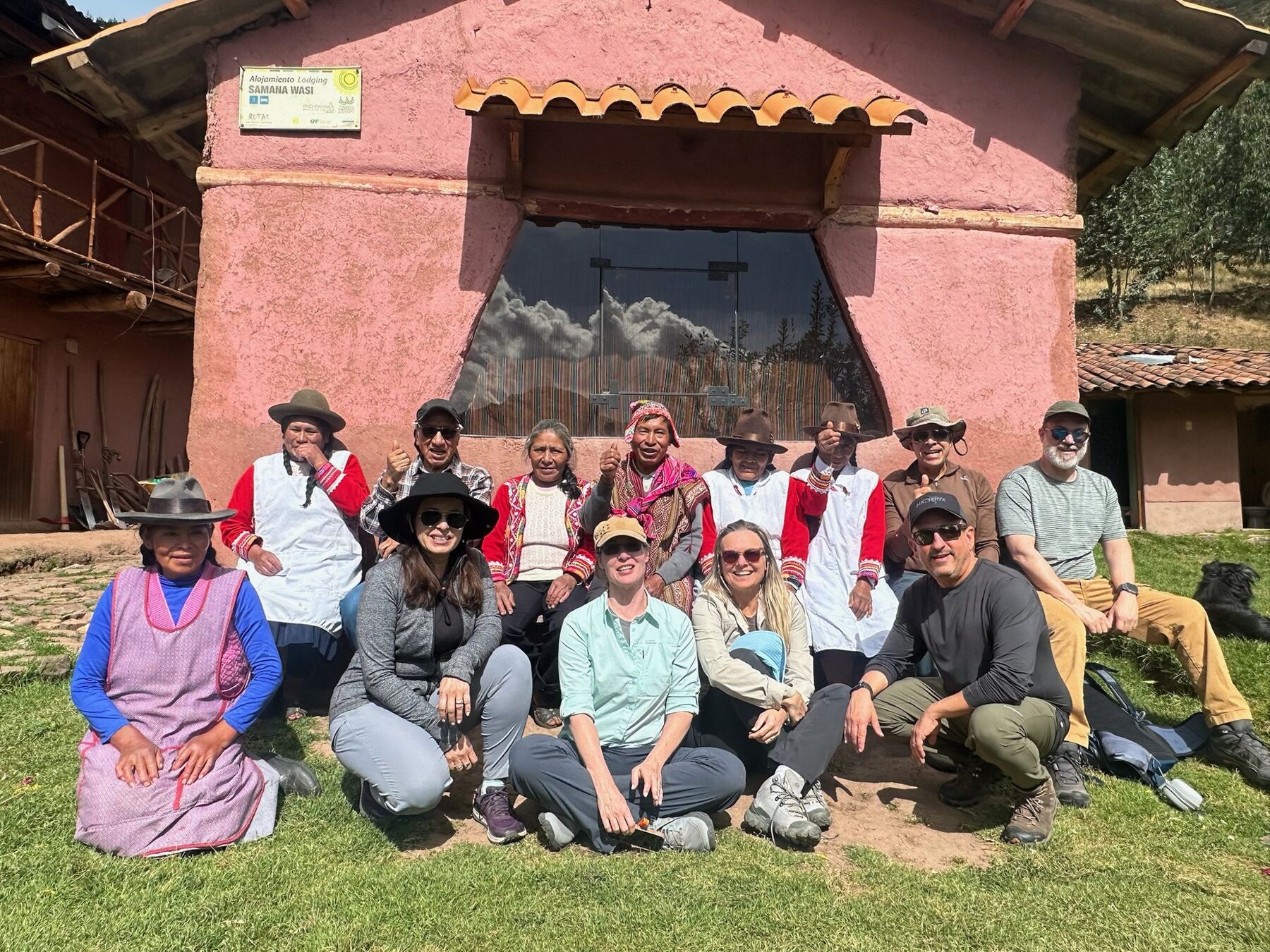 The travelers with community members in traditional dress in front of a pink adobe lodge