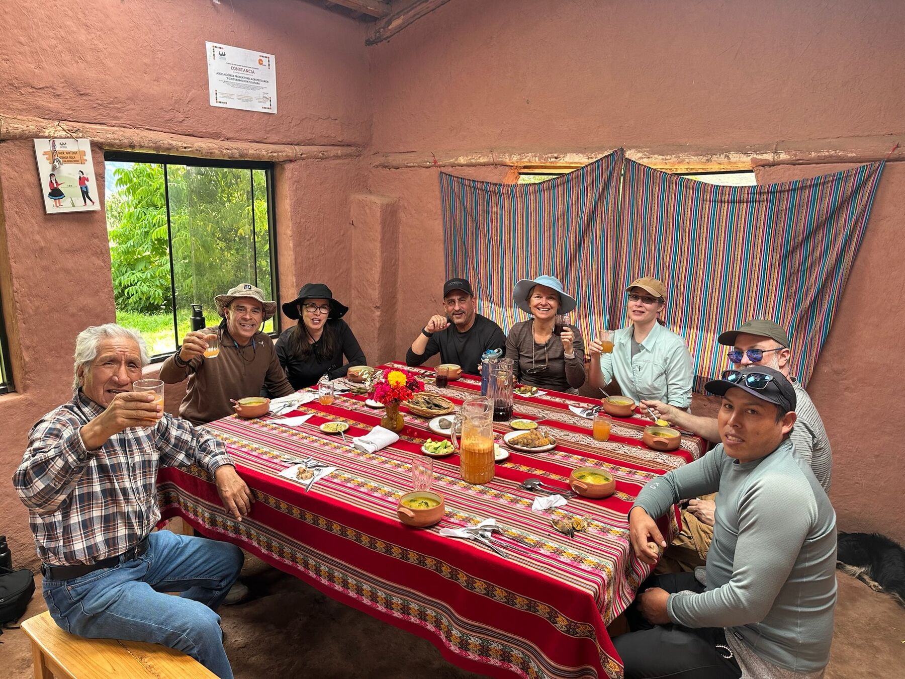 The travelers and their village hosts raising glasses around a table with a red woven cloth inside an adobe room