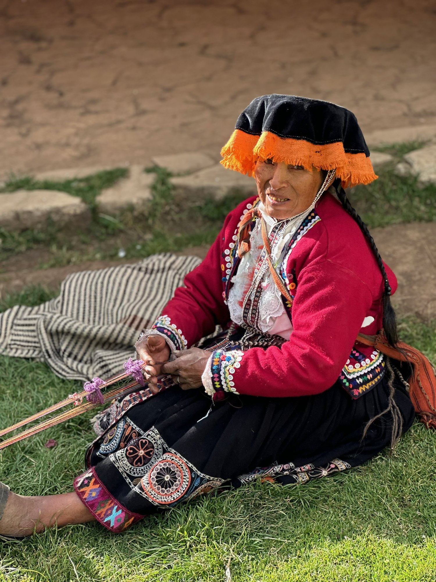 A woman in a red jacket and orange-trimmed black hat weaving a band by hand while seated on the grass