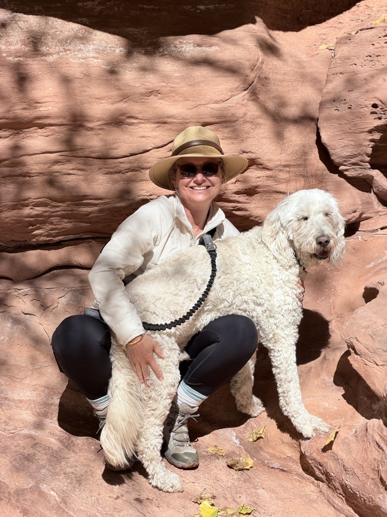 She-Stache crouching against a red sandstone wall off the Burr Trail with her arm around Pup-Stache, who is on a leash clipped to her waist