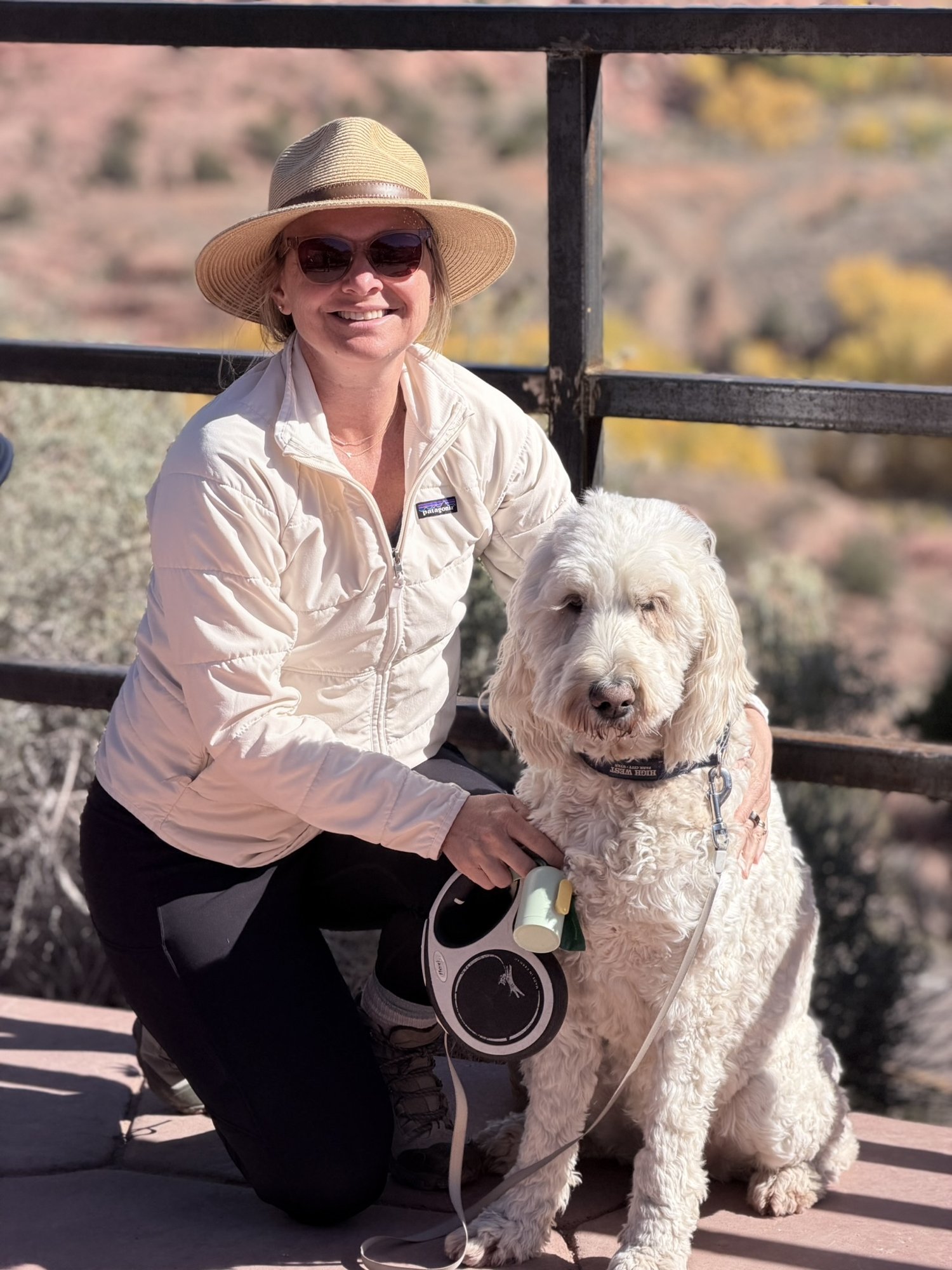 She-Stache kneeling beside Pup-Stache at a metal railing above the Escalante River canyon, yellow cottonwoods below