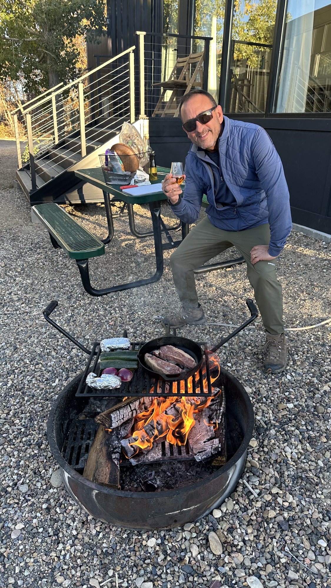 He-Stache sitting by a fire pit at Ofland Escalante with steaks on the grill and a drink in hand