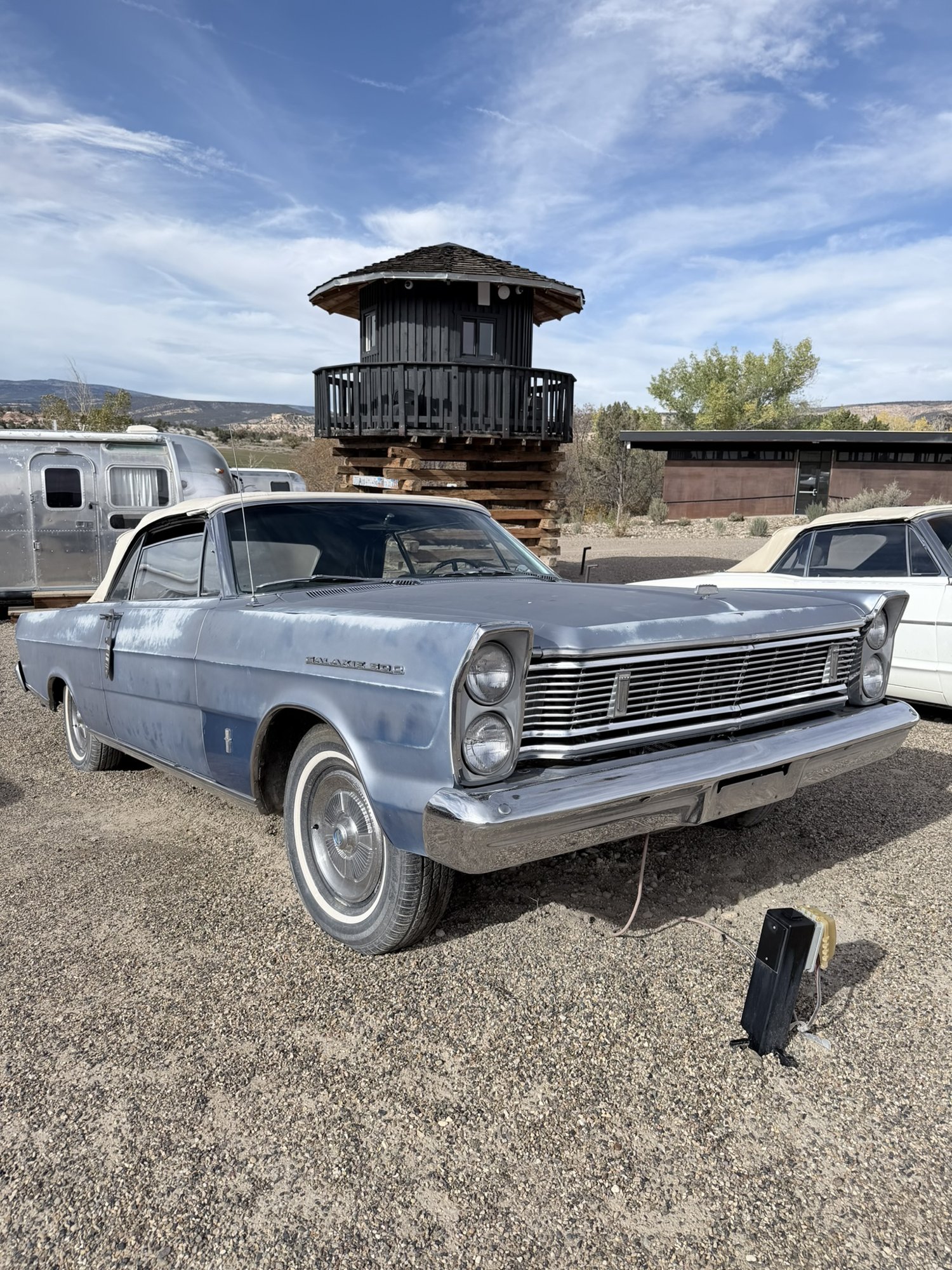 A faded blue vintage Ford Galaxie 500 convertible parked on gravel at Ofland Escalante, with an Airstream and a wooden lookout tower behind it