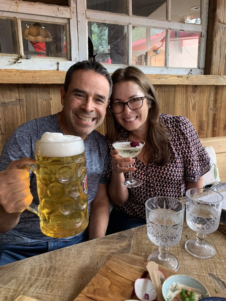 A grinning Scout-Stache raising a huge glass beer stein beside a smiling friend holding a coupe cocktail at Alpenrose