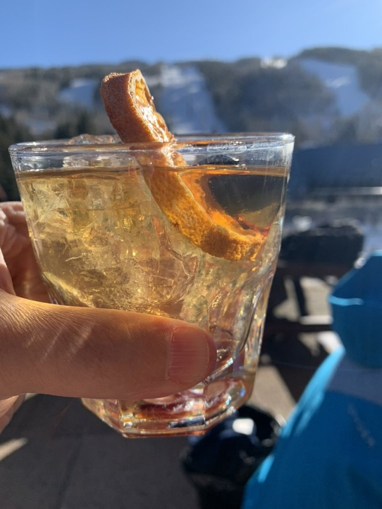 A hand holding an old fashioned with a dried orange peel, ski runs blurred behind it at the base of Vail