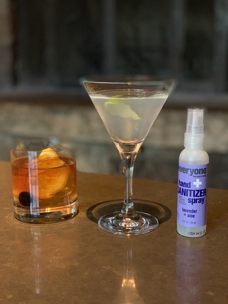 A martini and an old fashioned on a bar beside a small spray bottle of hand sanitizer