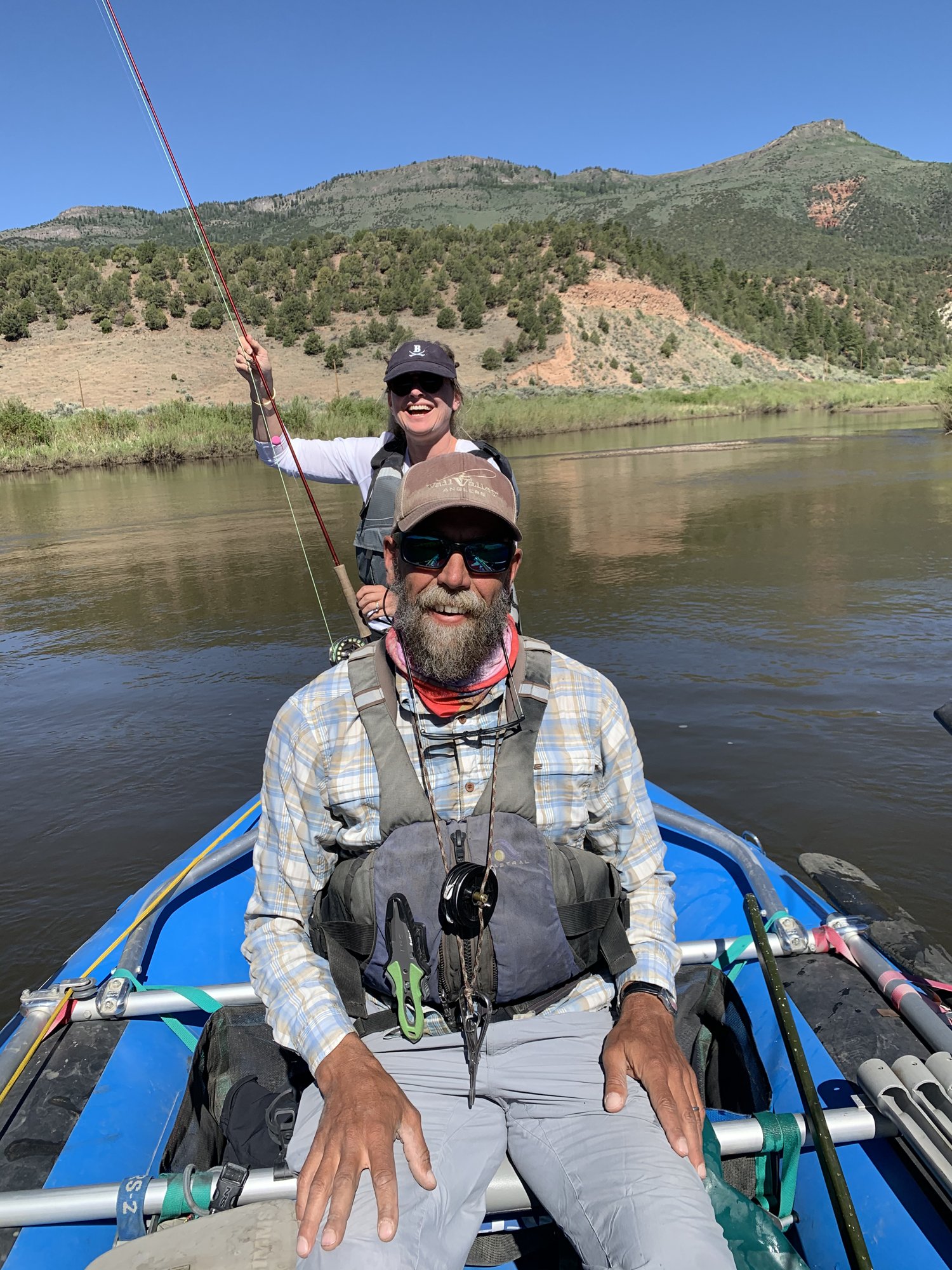 A fishing guide in a vest at the oars of a blue drift boat while She-Stache casts from the bow on the Colorado River