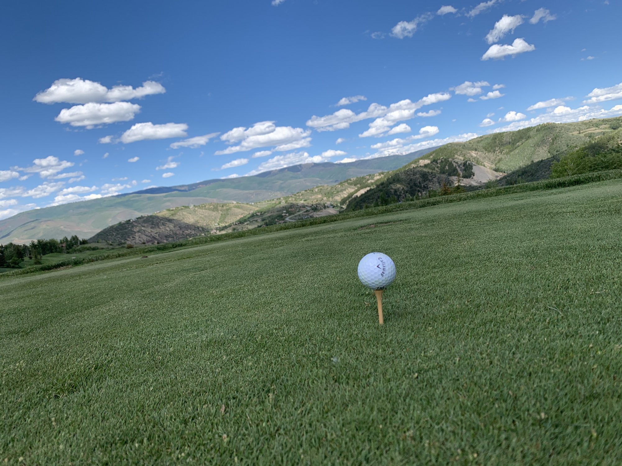 A golf ball on a tee on a bright green fairway sloping toward a mountain valley at Cordillera