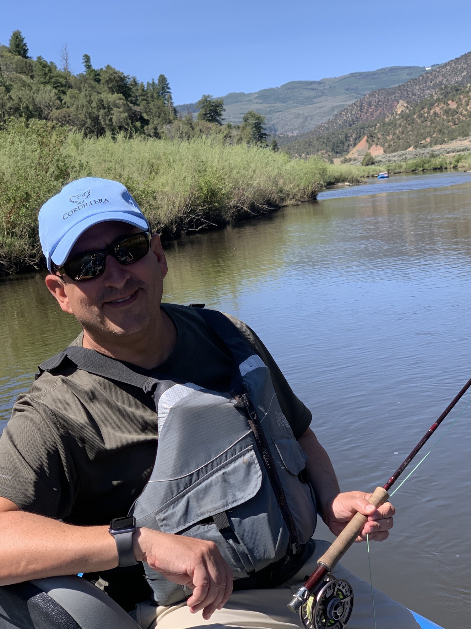 He-Stache in a light-blue cap and life vest holding a fly rod in a drift boat on a green river