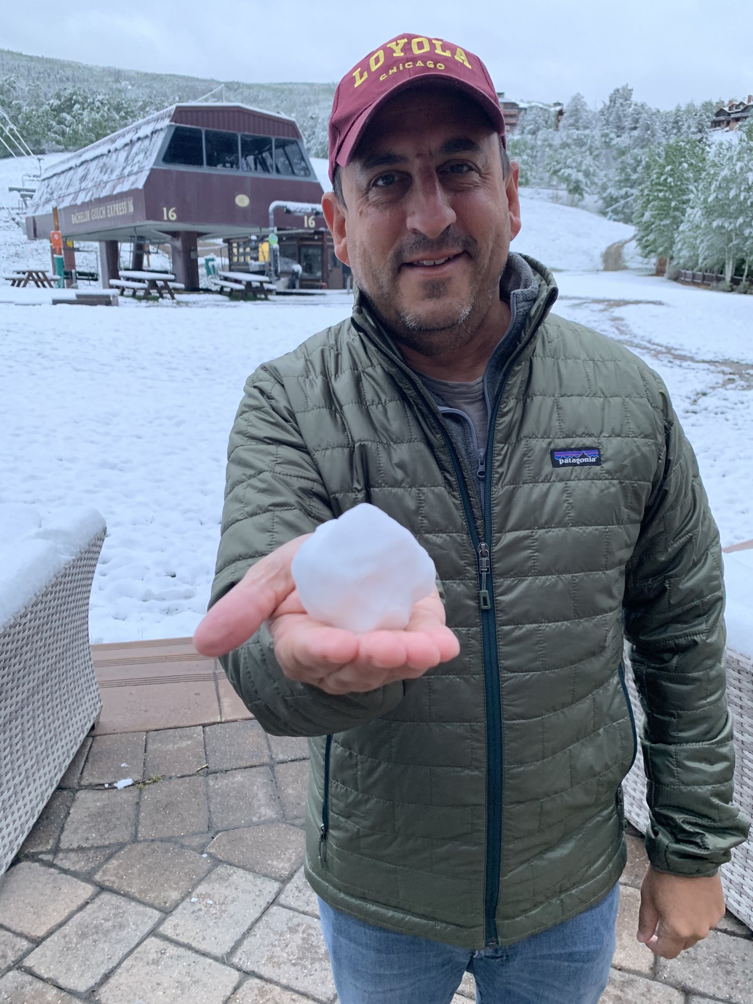 He-Stache in a maroon cap and green puffer jacket holding a snowball in front of a snowy lift station