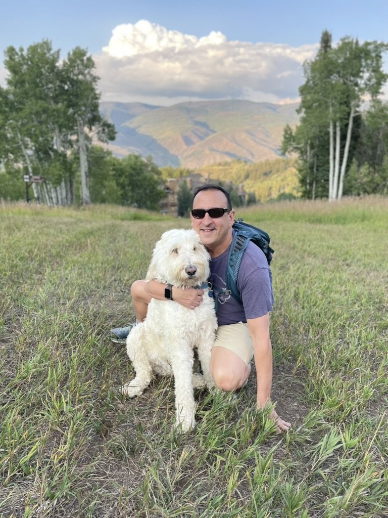 He-Stache in a hiking pack kneeling on a grassy ski run with his arm around Pup-Stache