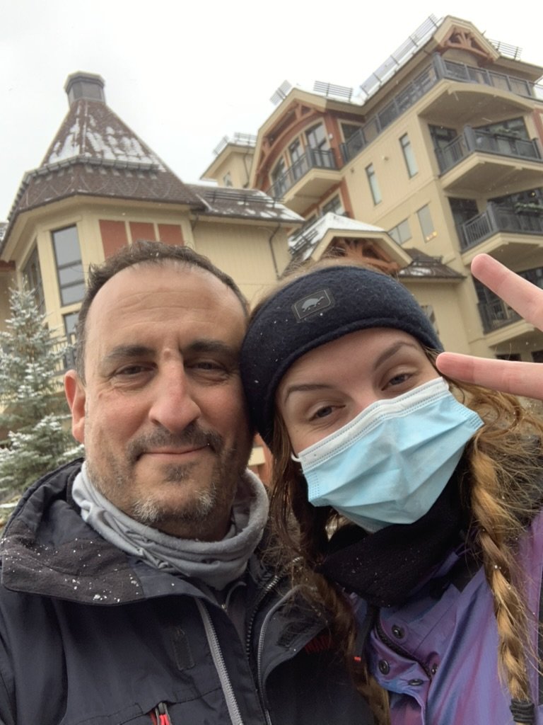 He-Stache with a short gray beard taking a snowy selfie with a Wee-Stache in a mask and braids, a lodge behind them