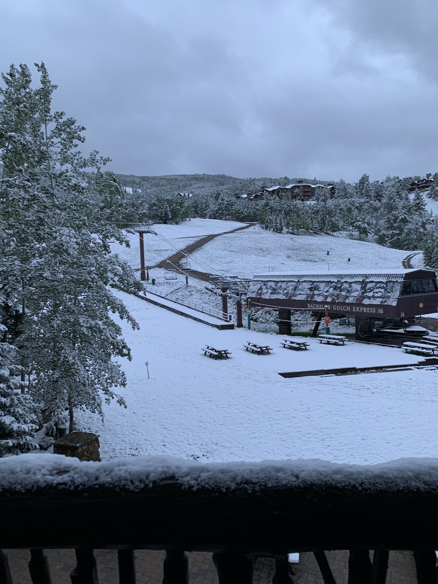 A snowy view of a chairlift station and picnic tables in June, framed by a snow-covered railing