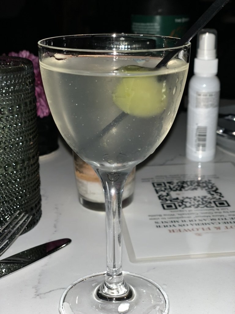 A martini with a skewered olive in a stemmed glass at Root & Flower, beside a menu card and a small spray bottle