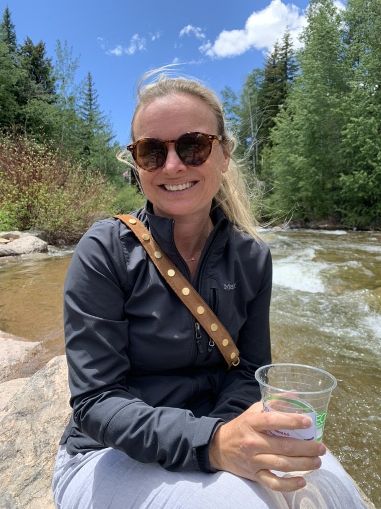 She-Stache in sunglasses and a dark jacket sitting on a boulder beside a rushing creek in Vail, holding a plastic cup