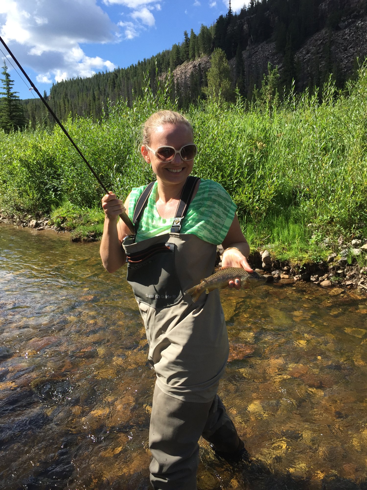 She-Stache in waders and sunglasses standing in a clear mountain creek near Red Cliff, holding up a small trout with her fly rod