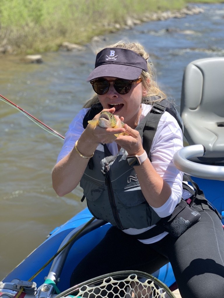 She-Stache in a visor and life vest jokingly kissing a small trout in a drift boat