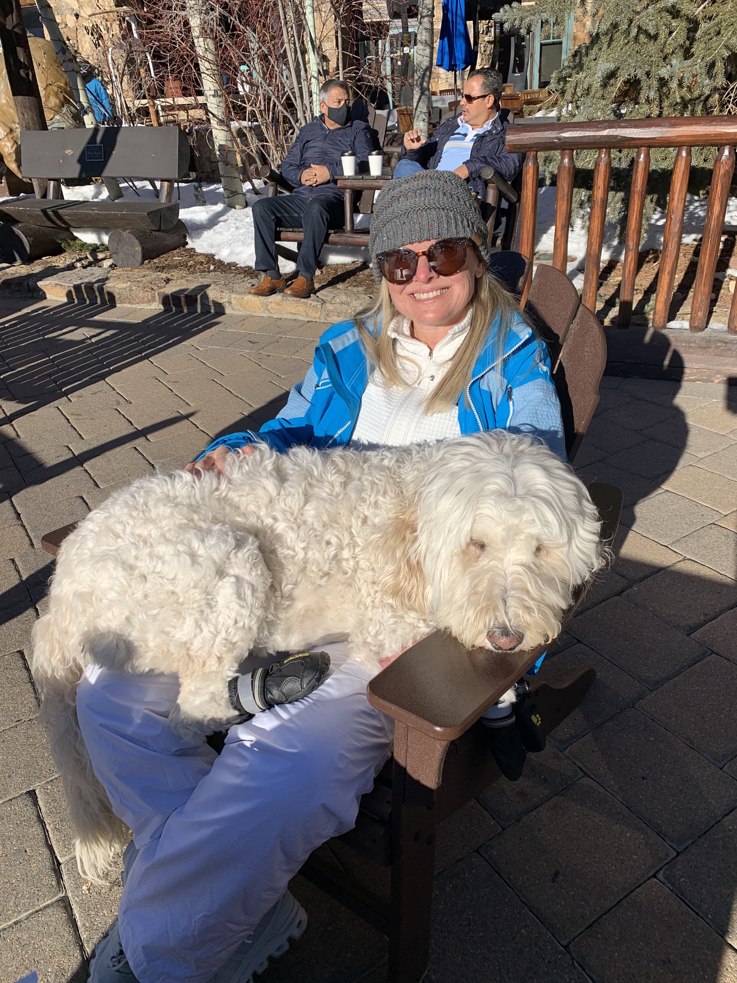 She-Stache in a blue ski jacket on a sunny patio with Pup-Stache sprawled across her lap