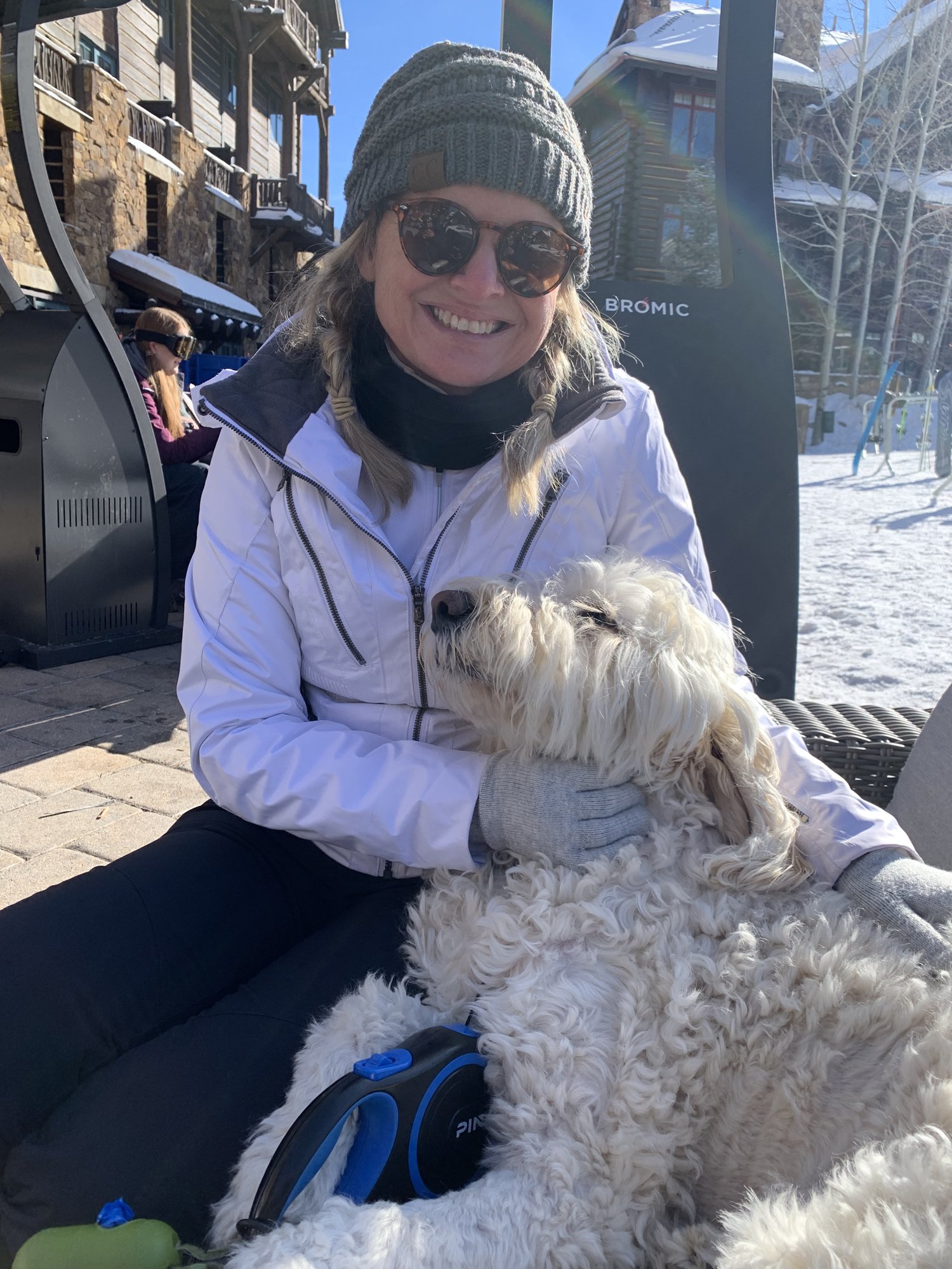 She-Stache in a white ski jacket and knit hat sitting outdoors at the Ritz-Carlton, Bachelor Gulch, with Pup-Stache snuggled across her lap