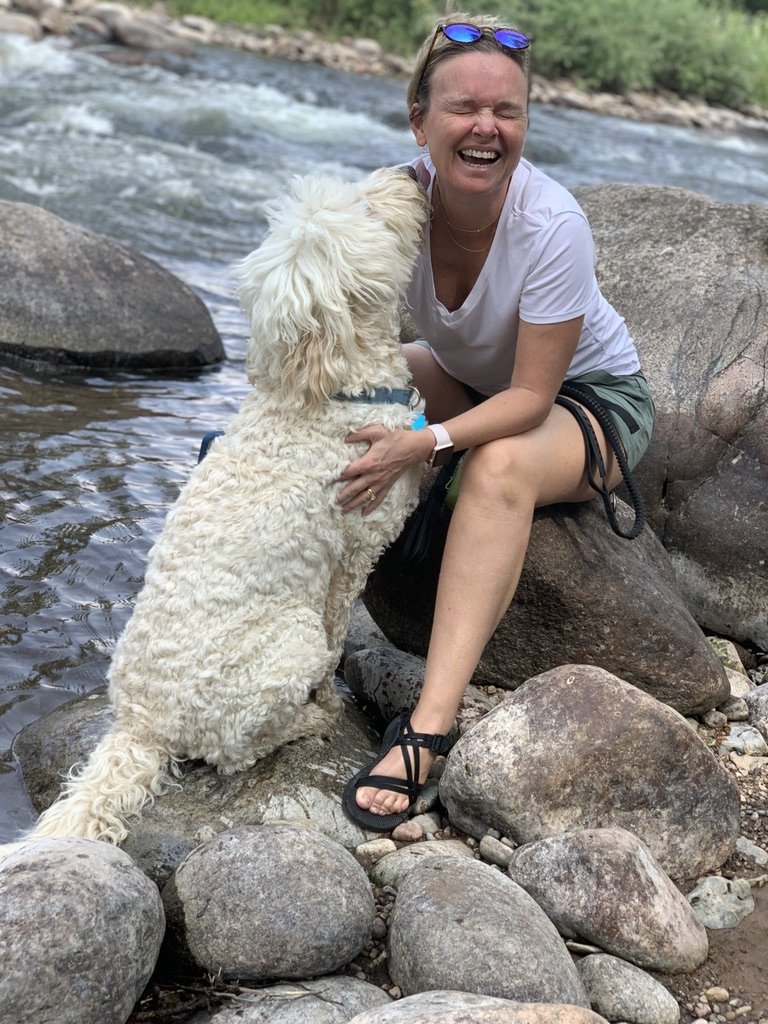 She-Stache sitting on river rocks while a wet Pup-Stache leans in for a hug