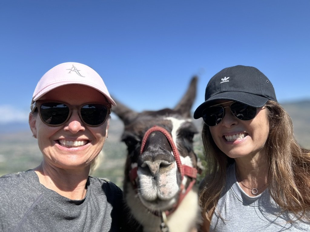She-Stache and a Scout-Stache in caps and sunglasses grinning beside a llama in a red halter