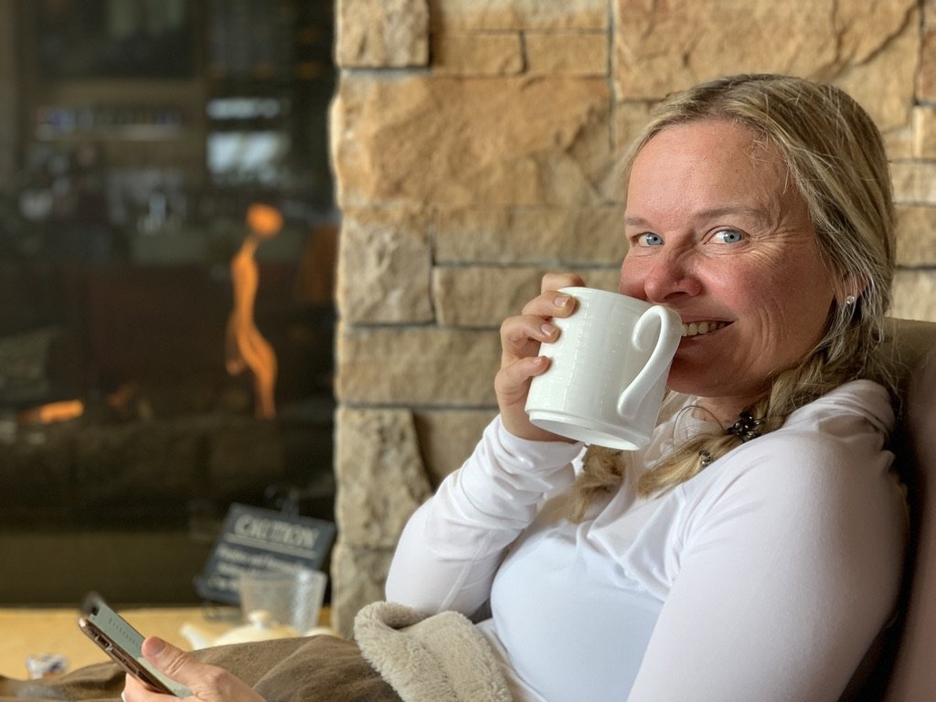 She-Stache in a white sweater sipping from a white mug beside a stone fireplace at The 10th