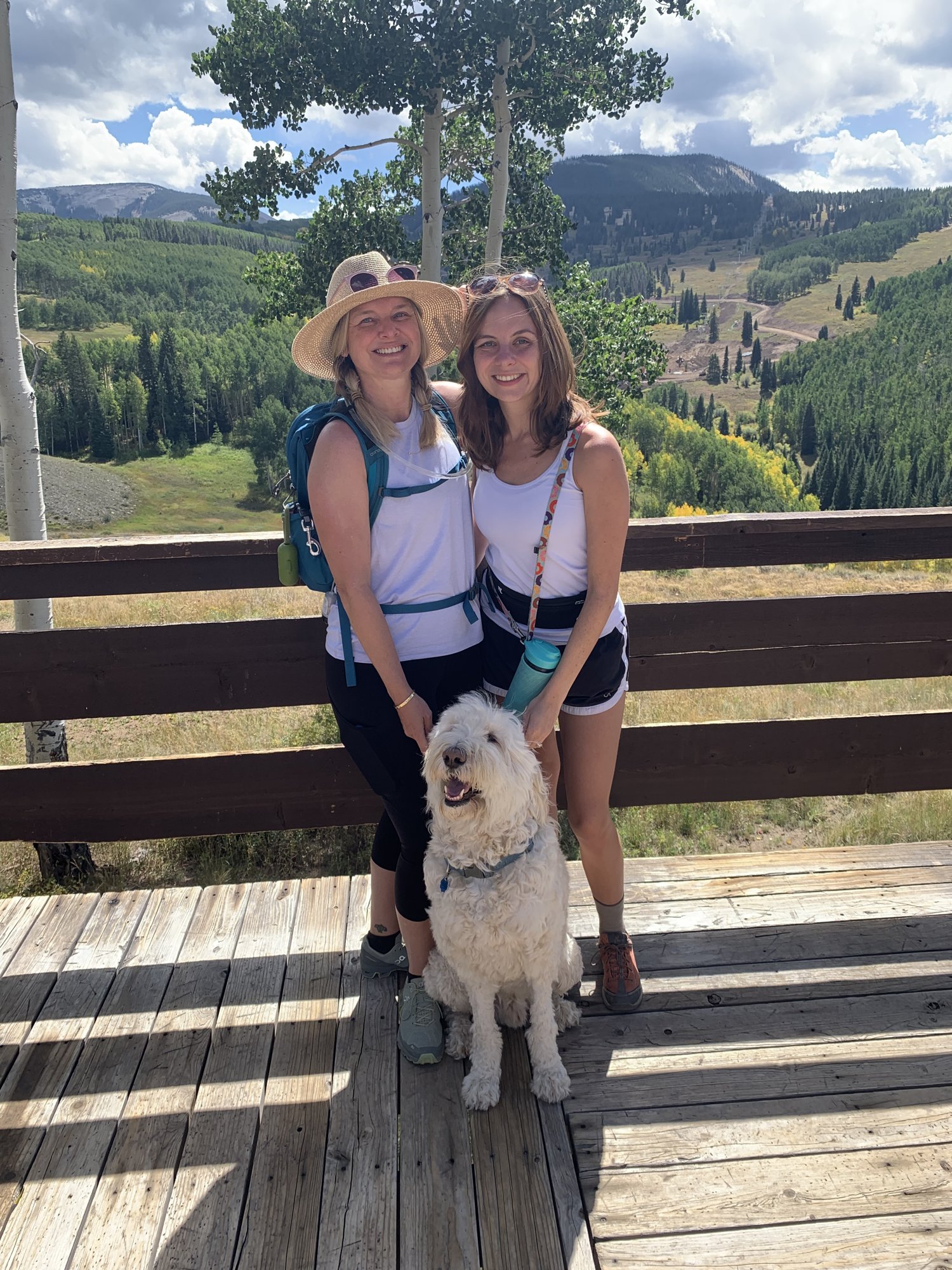 She-Stache and Wee-Stache 1 in hiking clothes sitting on a wooden deck with Pup-Stache, ski runs and hills behind them