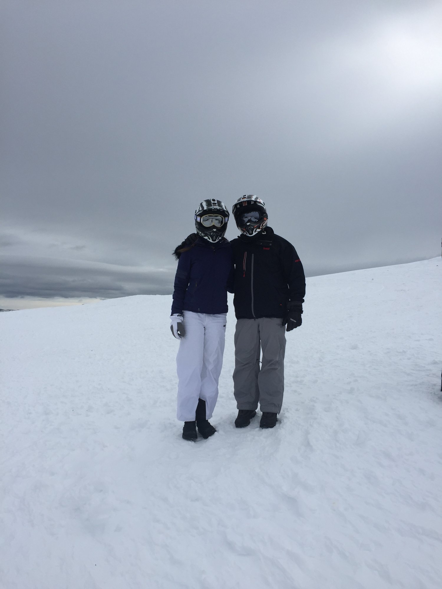 He-Stache and Wee-Stache 2 in snowmobile helmets and winter gear standing on a snowfield under a gray sky