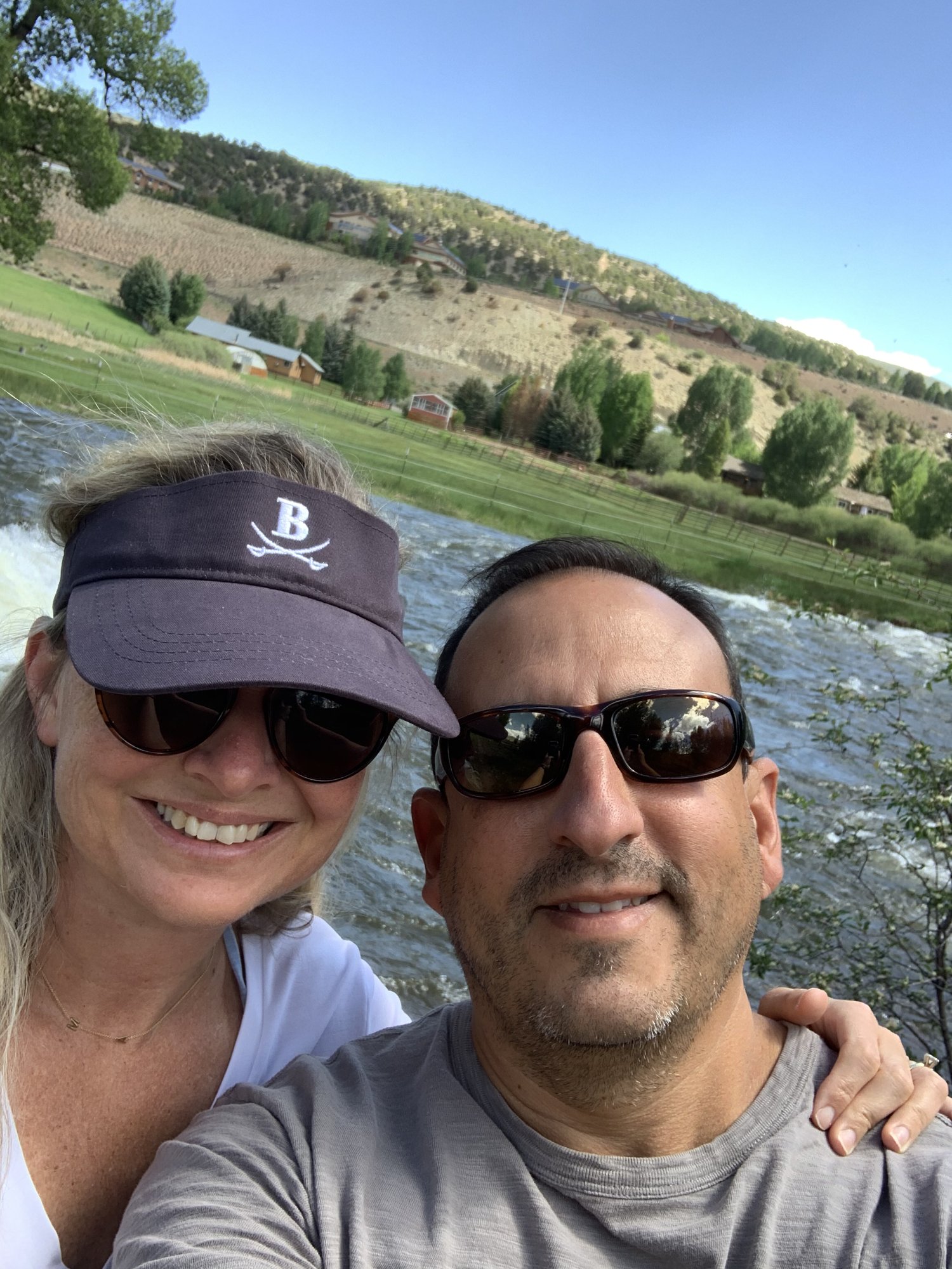 She-Stache in a visor and He-Stache in sunglasses taking a smiling selfie beside the rushing Eagle River in Edwards in June