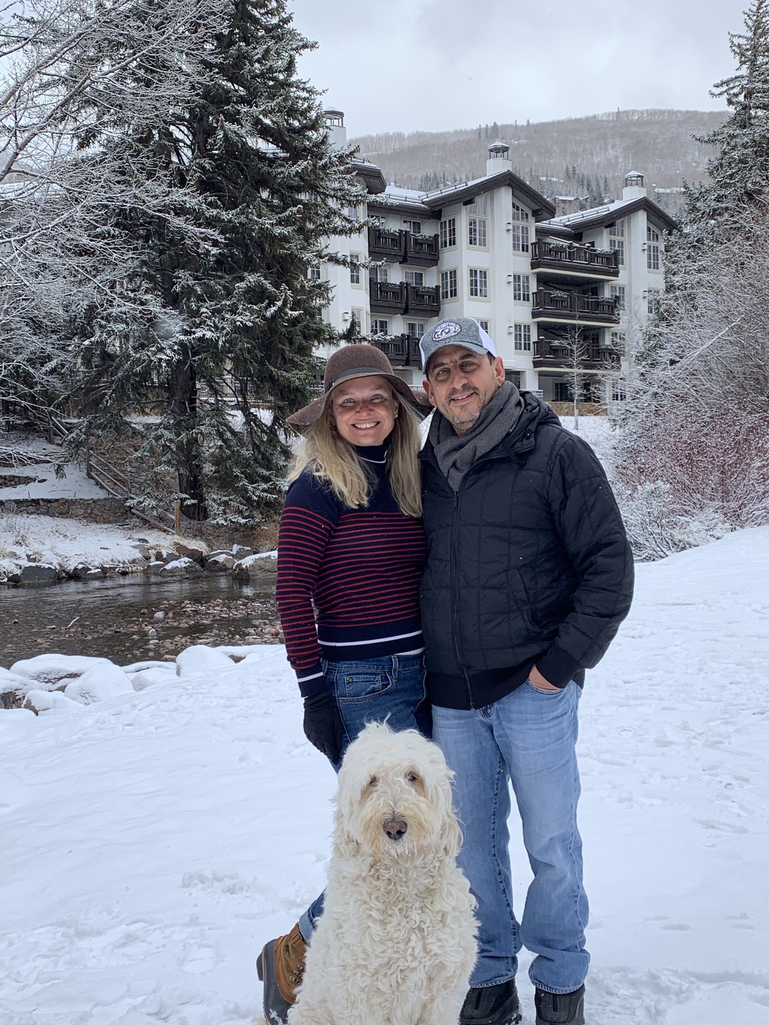 She-Stache and He-Stache in winter coats standing in fresh snow beside Gore Creek, Pup-Stache sitting in front of them, a snowy lodge behind