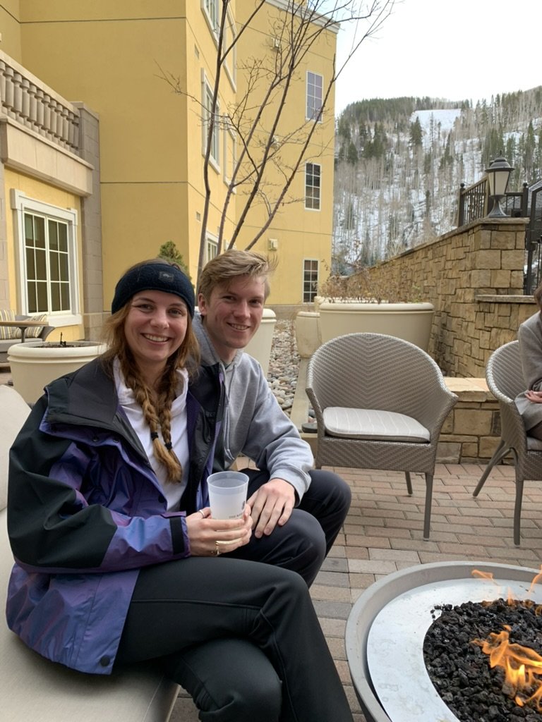 A Wee-Stache in a purple ski jacket and her now-husband in a gray hoodie beside an outdoor fire pit at the Ritz-Carlton Club, snowy slopes behind them
