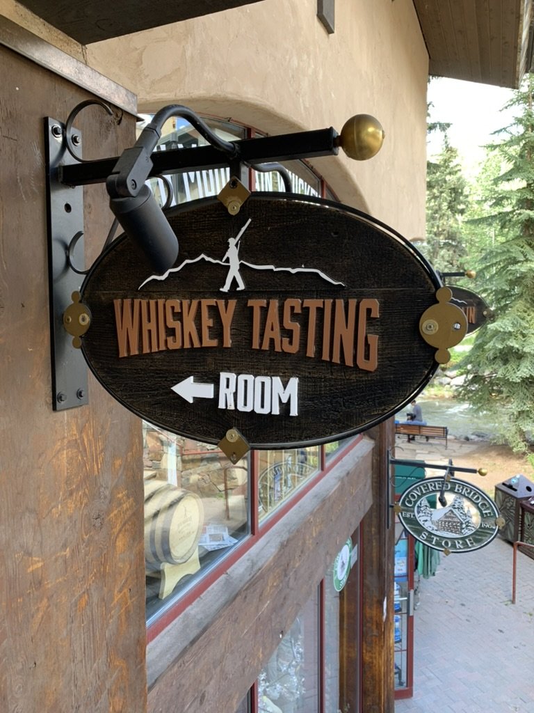 A carved wooden sign reading Whiskey Tasting Room with an arrow, hanging above a cobbled Vail Village street